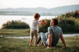 A father and son playing with a toy airplane while sitting on the grass near a lake with mountains in the background.