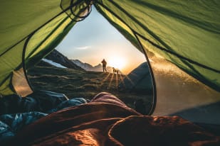 View from inside a tent showing a person walking in the distance at sunset with mountains in the background.