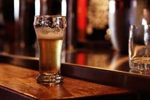 A half-filled glass of beer with froth sits on a wooden counter in a dimly-lit brewery bar, with a second empty glass in the background.