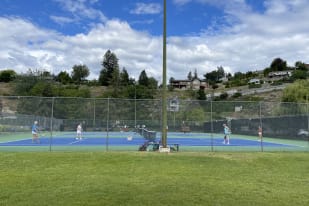 Tennis players on an outdoor court in Summerland under a partly cloudy sky with hills and houses in the background.
