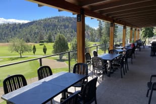Outdoor dining area with empty tables and chairs under a covered patio overlooking a golf course and scenic hills in Summerland.