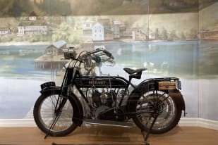 A black 1914 Hobart motorcycle displayed in a museum with a mural of a lakeside village in the background.