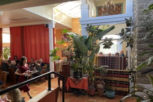 Cozy restaurant in Summerland with potted plants, red curtains, and diners enjoying meals, while a waitress in a red dress serves a table.