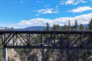 A black railway bridge stretches across a scenic gorge with a backdrop of forested hills and a partly cloudy sky in Summerland.