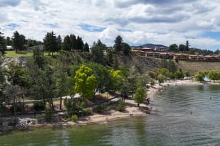A picturesque view of Summerland showing a beach, calm waters, treed hills, and houses on the hill with a partly cloudy sky overhead.