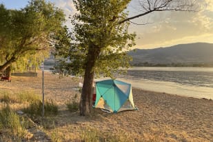 A colorful tent set up on a sandy beach by a calm lake, surrounded by trees, with mountains in the background during a sunset at Osoyoos.