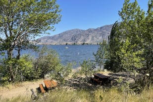 Scenic view of Osoyoos Lake with picnic tables under trees and mountain backdrop on a sunny day.
