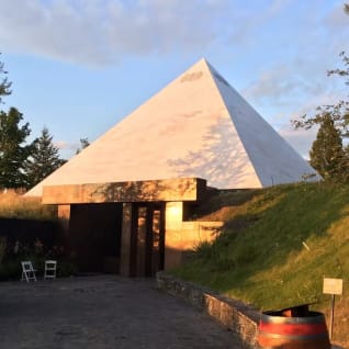 A building with a large triangular roof and a wooden barrel in front of it on a dirt path with a grassy hill in the background.