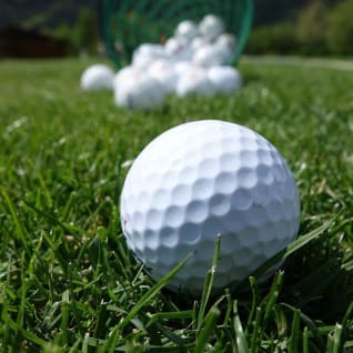 A close-up of a golf ball on grass with more golf balls and a basket in the background at The Golf Centre Practice Facility in Kelowna.