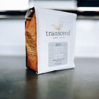 a bag of coffee sitting on top of a wooden table next to a bowl of fruit and coffee beans