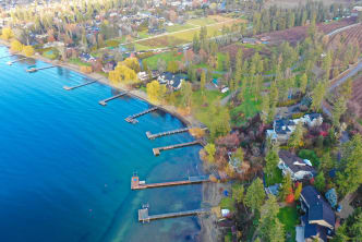 Aerial view of Kelowna featuring residential houses along the lakeshore with private docks extending into the blue water, surrounded by green trees and fields.