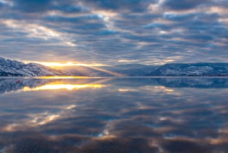 A serene sunset over a calm lake with surrounding snow-capped mountains under a cloudy sky in Kelowna.