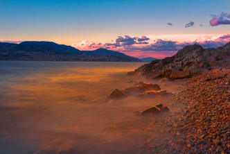 A rocky shoreline with water reflecting the vivid colors of a dramatic sunset over the mountains in Kelowna.