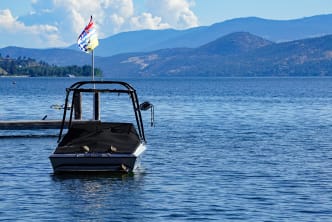 A boat with a covered bow and a flag docked on a lake, with mountains and a cloudy sky in the background.