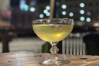 a glass of yellow liquid sitting on top of a wooden table in front of a building at night time