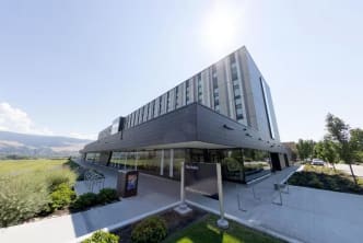 A modern multi-story building with a sign reading Nechako in front, surrounded by greenery and a clear sky in the background.