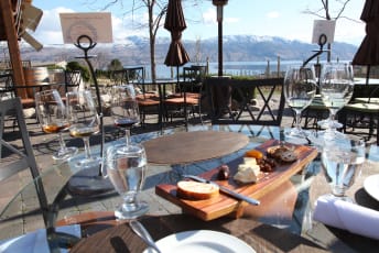An outdoor dining setup in Kelowna featuring wine glasses, water glasses, and a wooden board with assorted cheeses and bread on a glass table overlooking a scenic view of mountains and a lake.