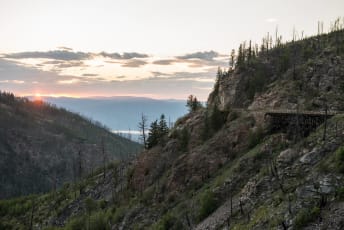 A scenic sunset over a mountainous landscape with a narrow bridge and sparse trees.