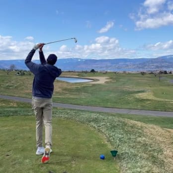 a man hitting a golf ball with a golf club on a golf course near a lake and mountains in the distance at The Harvest Golf Club, Kelowna.