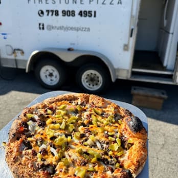 A freshly baked pizza with toppings of cheese, peppers, and meat in front of Krusty Joe's Firestone Pizza takeaway truck in Penticton, BC.