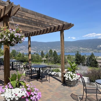 Outdoor seating area in Summerland with tables under a wooden pergola, hanging flower baskets, potted plants, and a scenic lake view with mountains in the background.