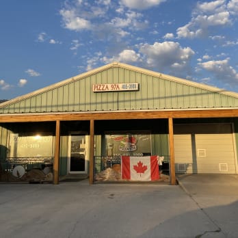 A pizza restaurant in Osoyoos adorned with Canadian flags during a sunny day, with a blue sky and scattered clouds above.