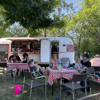People enjoying a meal at outdoor seating in Penticton, with a food truck and tables with red checkered tablecloths surrounded by lush greenery.