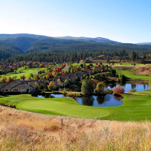 View of Gallagher's Canyon Pinnacle Course in Kelowna featuring green lawns, trees with autumn foliage, houses, ponds, and mountains in the background.