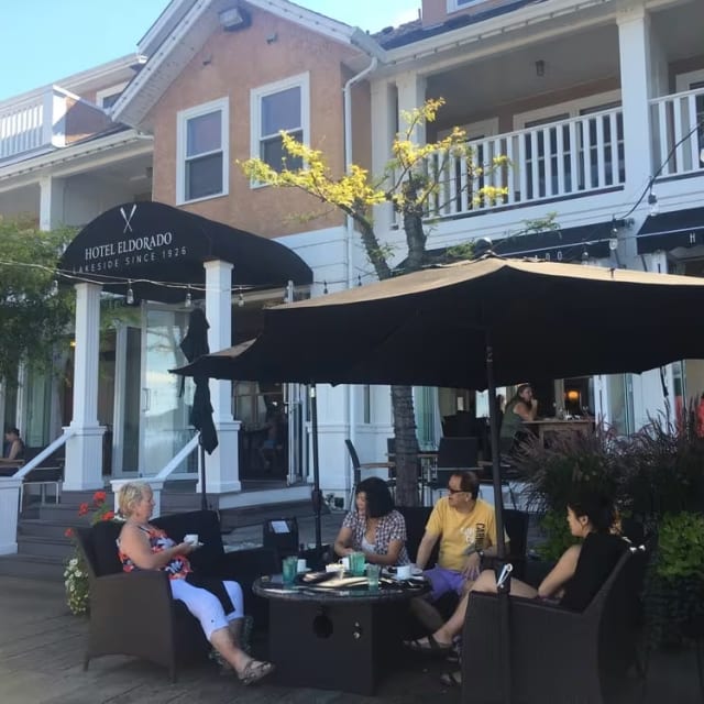 A group of people sitting around a table outside of the Hotel Eldorado building with umbrellas over it and people sitting at tables in front of the building in Kelowna.