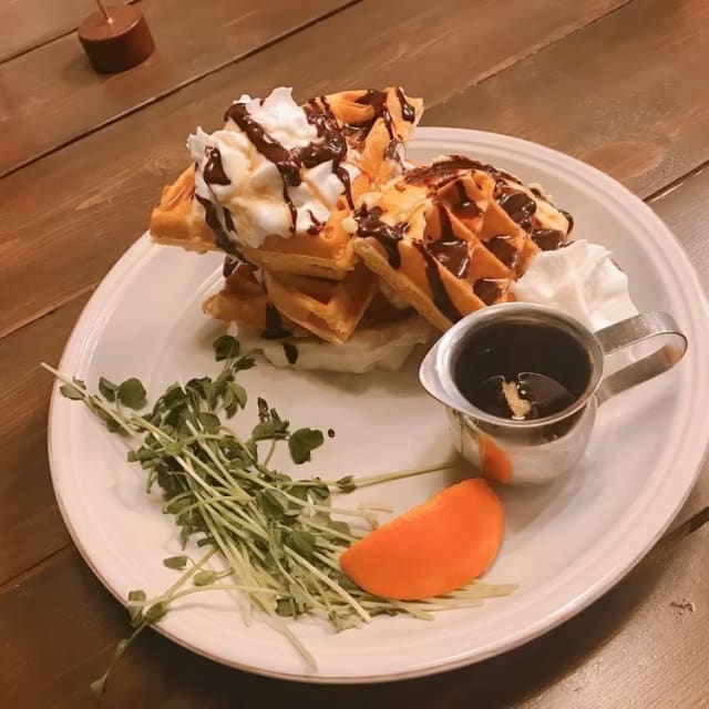 A plate of waffles topped with whipped cream and chocolate syrup, garnished with greens and a slice of apricot, next to a small pitcher of syrup, at Rustic Reel Brewing Co. in Kelowna.