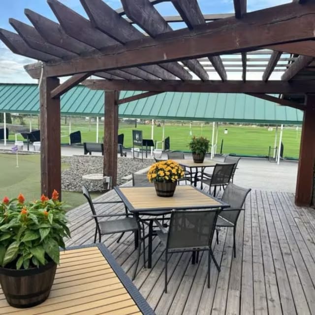 Outdoor seating area under a pergola at The Golf Centre Practice Facility in Kelowna, with a view of the green field in the background.