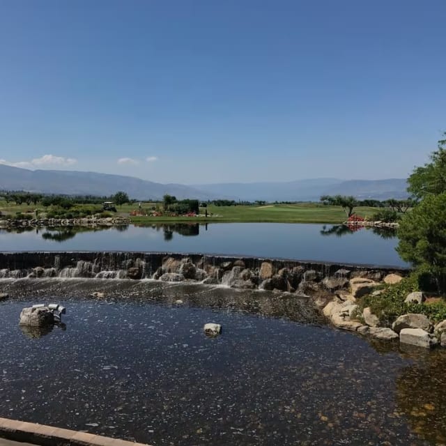 a pond with a waterfall in the middle of it and a golf course in the distance with mountains in the distance at The Harvest Golf Club in Kelowna.