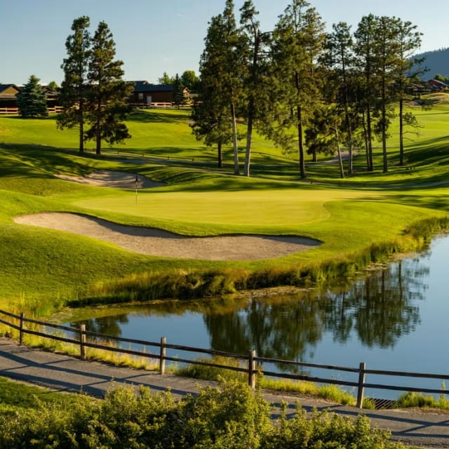 A scenic view of the Two Eagles Golf Course in West Kelowna, with a green surrounded by sand traps and a reflective water hazard under a clear sky.