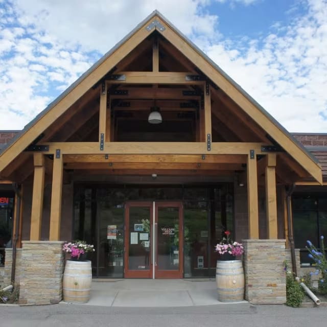 The entrance of Volcanic Hills Estate Winery in West Kelowna, featuring a wooden gabled roof, glass doors, and a stone façade, with flower pots and wine barrels on either side.