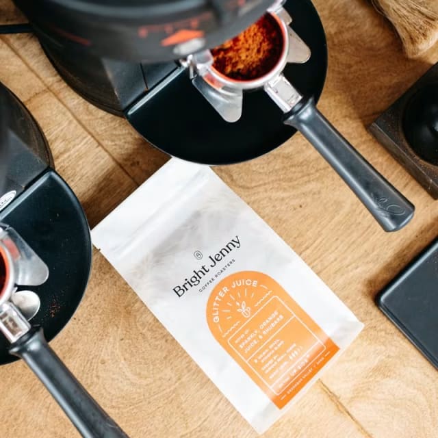 A coffee maker on a wooden table next to a bag labeled Bright Jenny Coffee Roasters and a cup filled with coffee grounds.