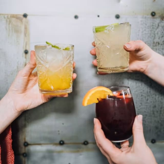 three people holding up glasses of different types of drinks in front of a metal wall with peeling paint on it