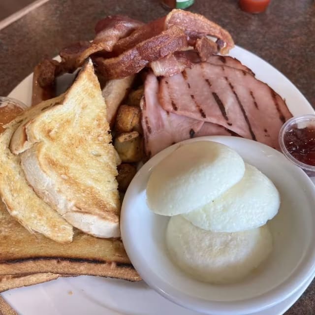 A breakfast plate from The Jammery in Lake Country featuring toast, bacon, ham, poached eggs, hash browns, and small portions of jam.