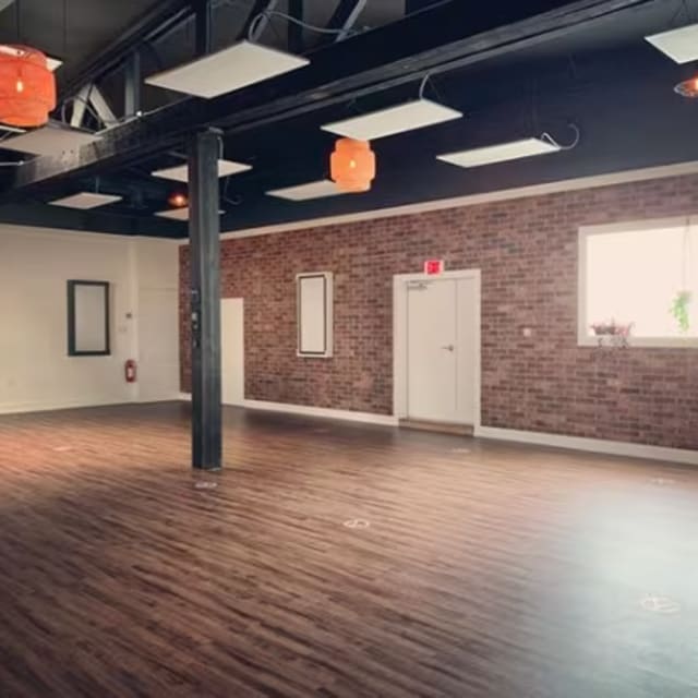 Spacious yoga studio in Kelowna with wooden flooring, exposed brick wall, ceiling lights, and natural light from a large window.