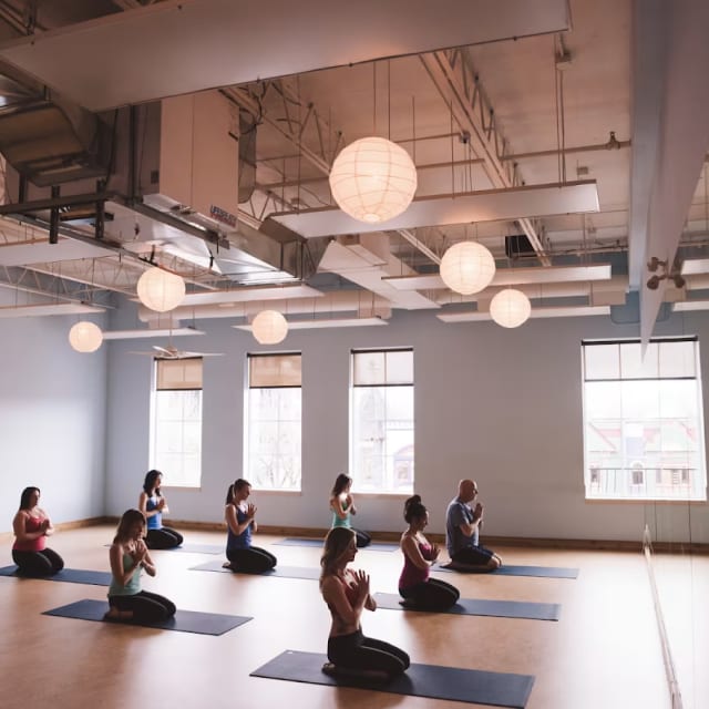 a group of people sitting on yoga mats in a large room with lights hanging from the ceiling and windows