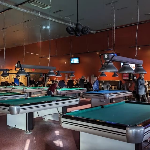 a group of people standing around a pool table in a room with a tv on the wall and a pool table in the middle of the room