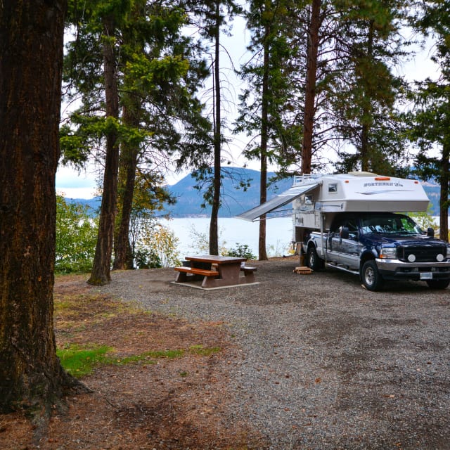 Camper truck parked at a scenic lakeside campsite with a picnic table surrounded by tall trees at Fintry Park in Vernon.