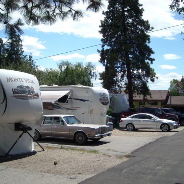 A view of Shady Lane RV & Trailer Park in Kelowna, featuring several RVs, trailers, and cars lined up under a clear blue sky with pine trees in the background.