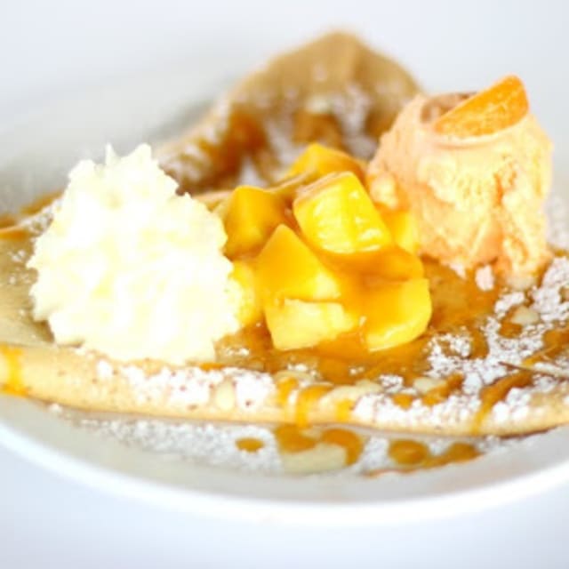 A crepe topped with whipped cream, mango pieces, mango ice cream, a citrus wedge, and drizzled with caramel.