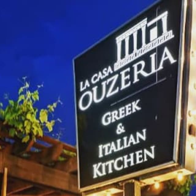 Neon-lit sign for La Casa Ouzeria Greek & Italian Kitchen, situated outdoors in a vibrant, leafy dining space.