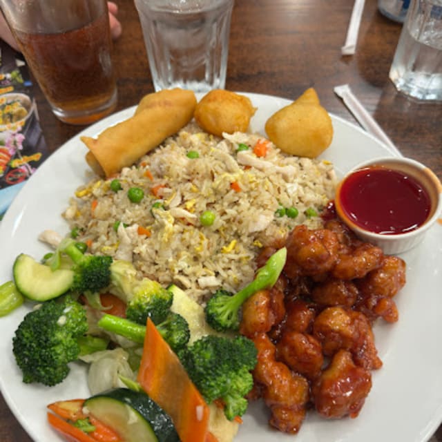 A plate of Asian cuisine with broccoli, carrots, zucchini, fried rice, spring rolls, fried chicken bites, and a small bowl of sauce, served with drinks in the background.