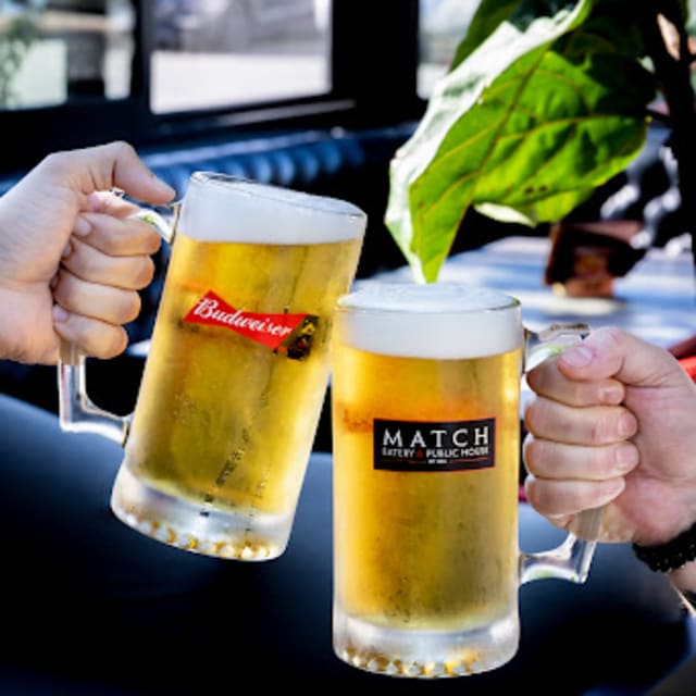 Two hands clinking large frosty beer mugs, one branded Budweiser and the other Match Eatery & Public House, in a casual setting with greenery in the background.