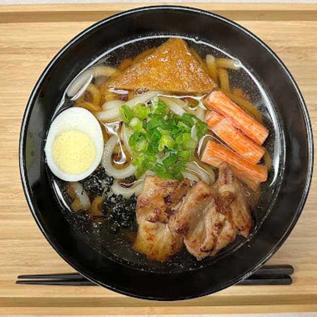 A bowl of Japanese donburi with noodles, boiled egg, crab sticks, grilled meat, green onions, and tofu on a wooden tray with chopsticks.