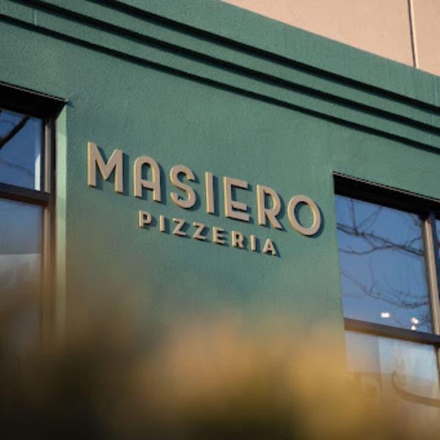 A green building facade with the words "MASIERO PIZZERIA" in gold letters, with tree reflections visible in the windows.