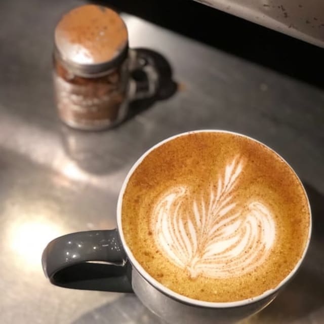 Close-up of a latte with intricate leaf latte art in a grey mug, placed on a metallic surface alongside a jar with a metal lid.
