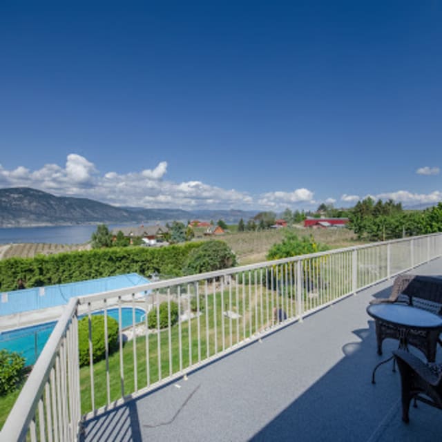 A spacious balcony at a bed and breakfast in Penticton overlooking a pool, lush greenery, and mountains under a clear blue sky.